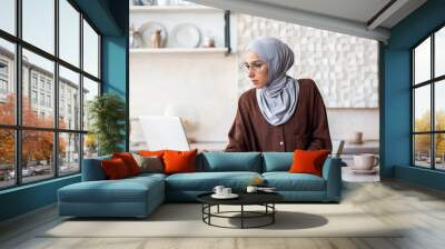 Young muslim woman in hijab freelancer working from home in kitchen. He sits focused on the laptop and makes notes in the notebook. Wall mural