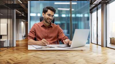 Young professional sitting at desk in modern office using laptop and taking notes. Man is focused on work, wearing stylish shirt, glasses, exuding confidence and motivation. Wall mural