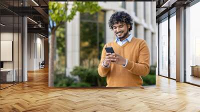 Young professional with curly hair using smartphone outdoors. Scene conveys connectivity, technology, and communication. Individual dressed in casual autumn attire, standing confidently near office Wall mural