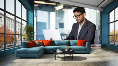 Young serious and focused businessman at workplace typing on computer, Arab man in business suit sitting at table thinking about solving technical work tasks. Wall mural