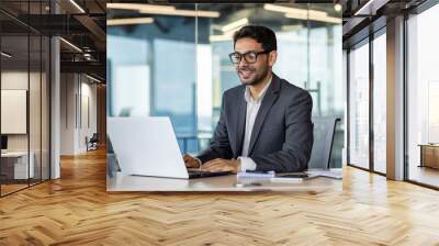 Young successful Arab businessman in business clothes works inside the office with a laptop, satisfied with the results, the boss in glasses types on the computer keyboard Wall mural