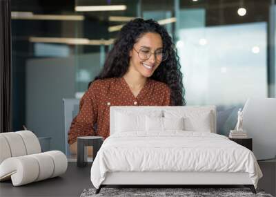 Young successful businesswoman behind paperwork, Hispanic woman working with documents and contracts inside modern office building, female worker using laptop, wearing glasses and curly hair smiling. Wall mural