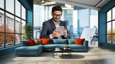 Young successful hispanic businessman at workplace using app on tablet computer, smiling man inside office building, working with laptop, wearing business suit, happy with achievement. Wall mural