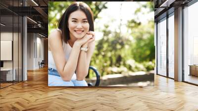 Beautiful asian woman sitting on bench and smiling. Modern girl resting in park on sunny summer day, looking happy Wall mural