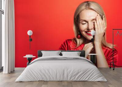 Beautiful middle aged woman with blond hair, stylish makeup, lauging and smiling, touching her flawless skin, standing over red background Wall mural