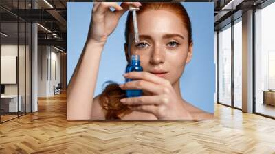 Beauty face of woman with freckles, natural clean and fresh glowing skin, apply anti-aging collagen serum. Hands holding serum with active botanical ingredients vitamin c in dropper, blue background Wall mural