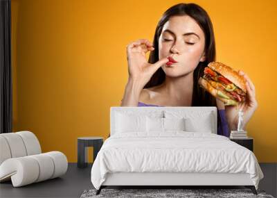 Brunette girl licks her finger, eats tasty hamburger. Woman orders takeaway food with delivery app, enjoys delicious burger, orange background Wall mural