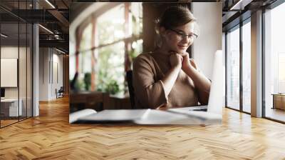 Girl being pleased with great job and positive result of work, sitting in quiet and cozy cafe, leaning head on hands and smiling broadly with satisfied expression at laptop, finishing project in time Wall mural