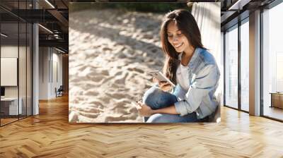 Girl writing post about beautiful sunny day on beach, edit pics from beach party as sit on hammock feet on sandy surface, smiling joyfully at smartphone screen, asking boyfriend take picture Wall mural