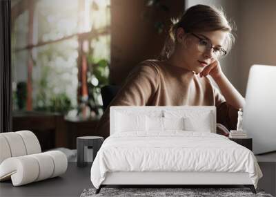 Good-looking young female freelancer tutor sitting in cozy cafe, leaning on hand while checking schedule via laptop, gazing at screen with focused expression, working, preparing material, making notes Wall mural