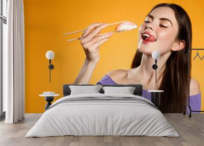 Hungry beautiful girl eats sushi with chopsticks, enjoys fresh nigiri in asian restaurant, orange background Wall mural