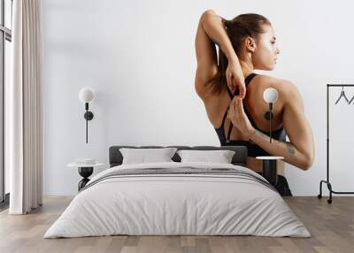 Rear view of fit and sporty fitness woman stretching arms and back before workout exercises. Woman back in black sport clothing standing on white background, training concept Wall mural