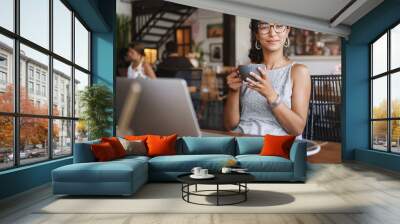 Relaxation, urban people and education concept. Charming young female freelancer, student having break from work, order coffee, holding cup and enjoy cappuccino, smiling pleased, using laptop Wall mural