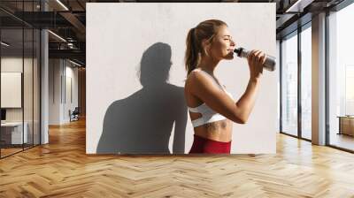 Satisfied female runner drinking water and smiling after workout. Woman athlete taking break while jogging outdoors, standing by concrete wall Wall mural