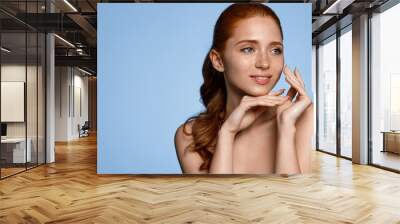 Skin care. Woman with beauty face touching healthy facial skin portrait. Beautiful smiling redhead girl model with natural makeup touching glowing hydrated skin on blue background closeup Wall mural