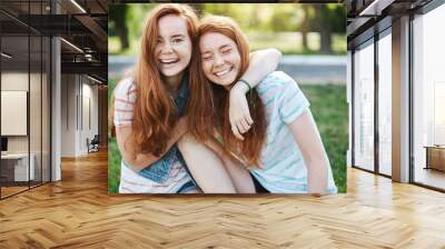 Wanna hug her forever. Portrait of happy and carefree two twin sisters with natural red hair and freckles, laughing out loud and cuddling, fooling around while resting in park on fresh green grass Wall mural