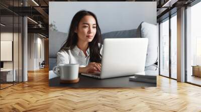 Young ambitious asian girl working remote from home, looking at laptop screen and smiling. Woman checking mail or researching while telecommuting, sitting on floor at her apartment Wall mural