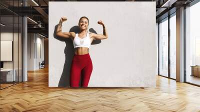 Young healthy woman doing fitness outdoors. Female athlete showing muscles, flex biceps and smiling satisfied, feeling strong after morning exercises Wall mural