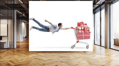 Full length shot of a casual young man flying and holding a shopping cart with christmas presents Wall mural