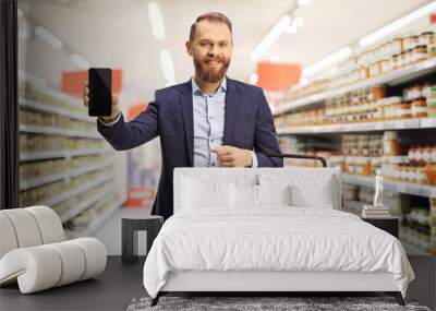 Smiling young man holding a shopping basket and showing a mobile phone Wall mural