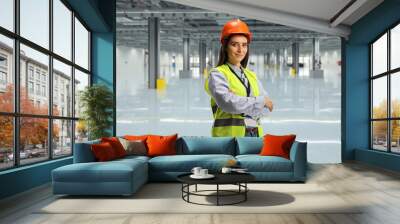 Young female engineer wearing reflective vest and helmet and standing in an empty factory Wall mural