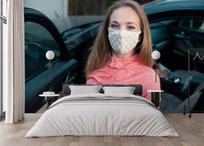 Young woman in fabric face mask next to her car. Many countries recommend citizens cover their faces during the world coronavirus pandemic. Wall mural