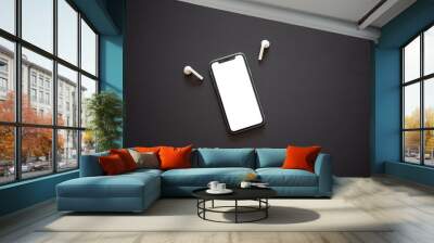 Modern smartphone with the turned on white screen and two white wireless headphones on a dark (black) background. Modern and minimalistic concept. Wall mural