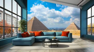 Two majestic pyramids stand tall against a backdrop of a clear blue sky and fluffy white clouds. The desert stretches out in front of them, leading to a city in the distance. Wall mural