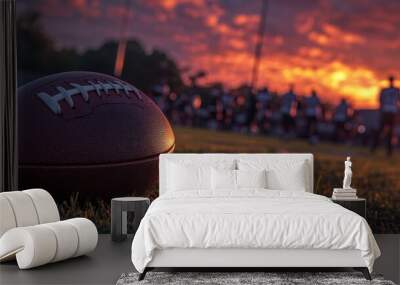 Close-up of a football on a grassy field with a blurry sunset and players in the background. Wall mural