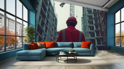 Construction worker in red uniform and hardhat stands between two buildings under construction. Wall mural