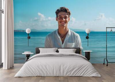 Smiling young man standing on a sunny beach with clear blue ocean and distant mountains Wall mural