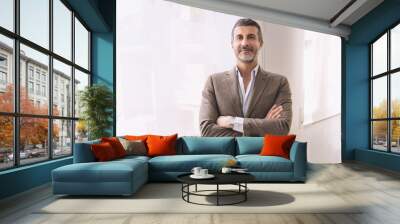 Confident Businessman in Bright Office Setting - Portrait of mature businessman with crossed arms, standing in white office illuminated by large window, in his forties, looking at camera Wall mural