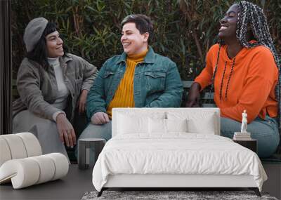 Diverse Friends Relaxing in Park - three young friends, a brunette woman, a non-binary plus size person, and an African woman with dreadlocks, casually dressed, sitting on a park bench. Wall mural