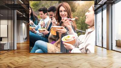 Gathering of young people eating take away organic food in plastic-free bowls and cutlery together  in the park. Focus on the second from right smiling and talking. Wall mural