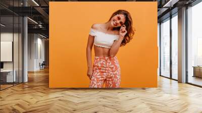 Active girl in orange pants with white leaves smiling radiantly, looking at camera through rose-colored glasses in cute studio Wall mural