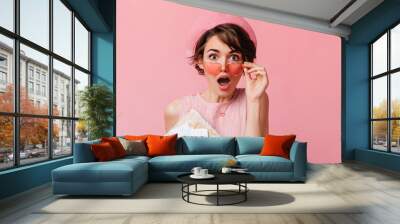 Adorable brown-haired woman with tickets touching glasses. Front view of shocked female tourist isolated on pink background. Wall mural