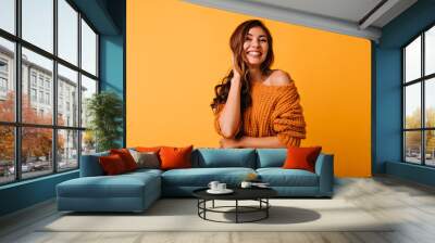 Adorable woman in orange attire touching her brown wavy hair. Laughing blithesome girl posing on yellow background. Wall mural