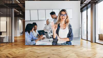 African students with serious face expression preparing for seminar while blonde girl posing with okay sign. Young female businesswoman having fun in office after brainstorm with colleagues. Wall mural