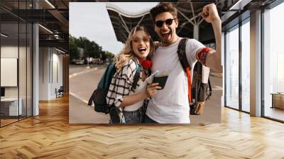 Attractive blonde woman in sunglasses and man in white tee smiles and takes selfie near airport. Portrait of happy travelers with backpacks. Wall mural