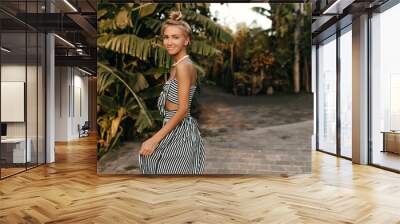 Attractive happy girl in striped stylish dress walks in tropical park. Charming blonde tanned woman in summer outfit enjoys sunny weather. Wall mural