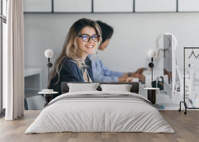 Attractive laughing freelancer girl posing with cup of coffee at her workplace. Chinese student in blue shirt works with document in campus with blonde friend in glasses. Wall mural