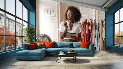 Attractive young woman in stylish white blouse and khaki shorts looks away, smiles, holds pen and computer tablet in cozy room. Wall mural