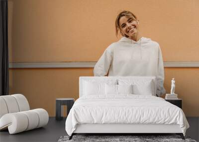 Beautiful caucasian young girl smiling with teeth looking at camera on background with place for text. Swarthy blonde beauty wears white warm tracksuit. Good mood, fashion trends Wall mural