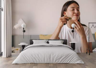 Beautiful young caucasian woman enjoys morning routine with face massager at home. Brunette hair girl sitting on bed in morning. Concept scraper, massage. Wall mural