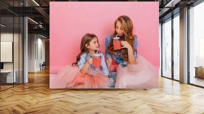 Beautiful young mom and curly daughter in same lush skirts drinking strawberry juice on pink background. Charming stylish woman and her little sister sitting on the floor with glasses of pink soda Wall mural