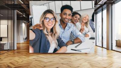 Black office worker in checkered shirt embracing blonde secretary girl while she making selfie. Young managers of international company having fun during meeting. Wall mural