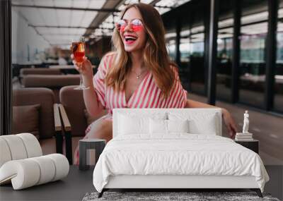 Blissful fair-haired girl spending summer day in restaurant. Happy european woman wears striped dress drinking champagne and smiling. Wall mural