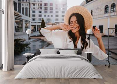 Blissful woman in summer hat posing in city. Outdoor photo of blithesome brunette girl in white blouse walking around town. Wall mural