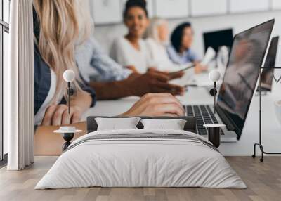 Blonde woman with elegant hairstyle typing text on keyboard in office. Indoor portrait of international employees with secretary using laptop on foreground. Wall mural