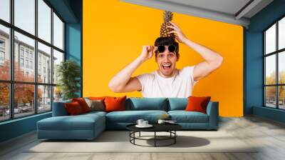 Brown-eyed naughty man takes off his sunglasses and holds pineapple on his head Wall mural